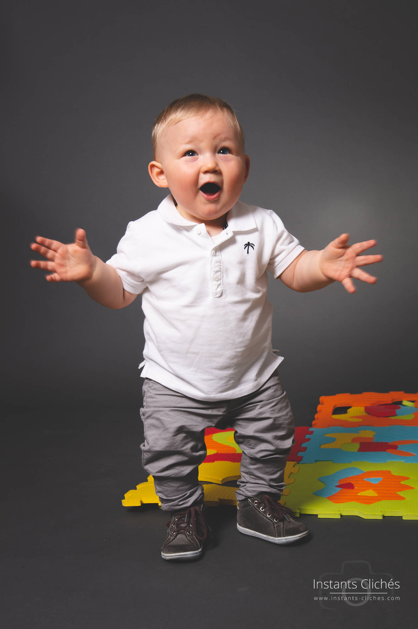 Bébé souriant avec bras ouverts sur fond gris.